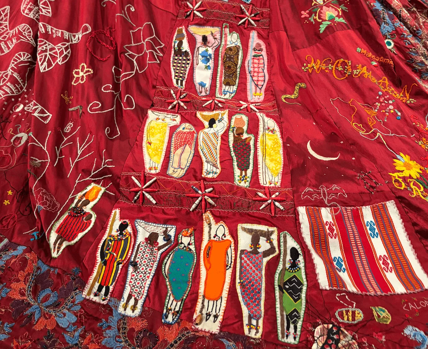 Detail of a red textile artwork with embroidered figures and flowers.